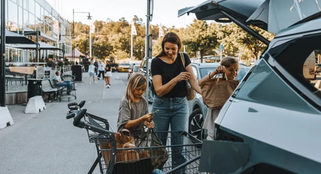 Children helping mom put groceries in the back of a vehicle. Family is in the parking lot loading area of a grocery shopping center.  