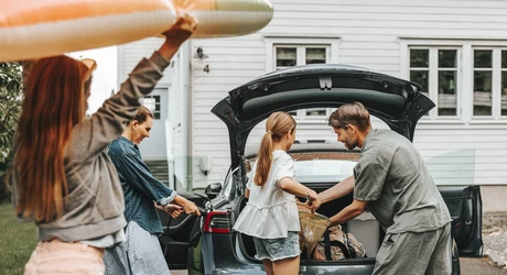 Woman charges auto in driveway, while family loads up trunk for a picnic beach day. Everyone is wearing lighter-weight summer clothing. 