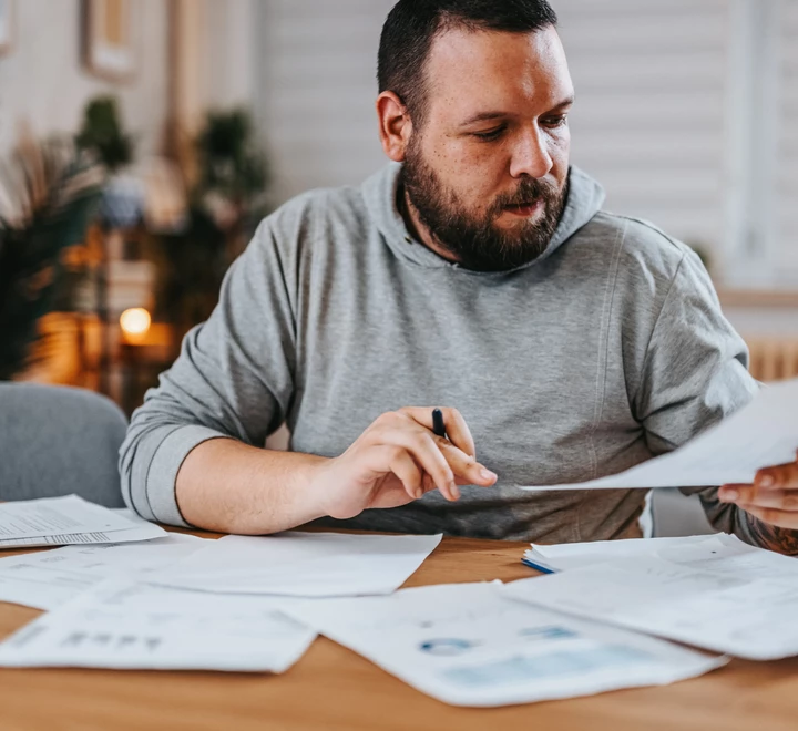 Man looking over his finances at home. Papers are on the table. The man has a pen in one hand and a piece of paper he’s reviewing in another hand.  
