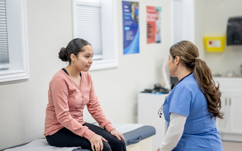 A young female teenager sits up on an exam table at the doctors during a routine check-up. She is dressed casually and her female nurse of Hispanic decent, is seated in front of her as they talk.