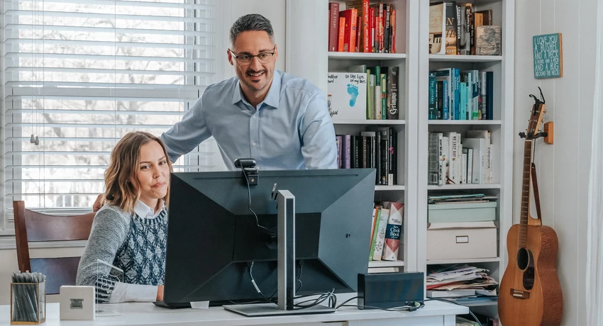 Two adults at a desk working on a computer in a bright home office, with one person seated and the other standing nearby. Behind them are bookshelves filled with books and decor, and a guitar rests against the wall next to the shelving. 