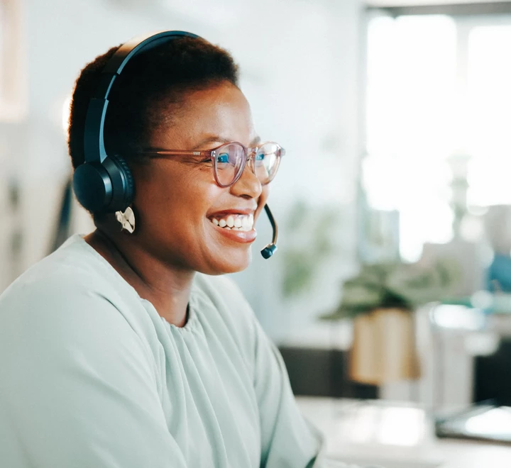 Person wearing a headset seated at a desk in an office, with a computer and shelves in the background. 