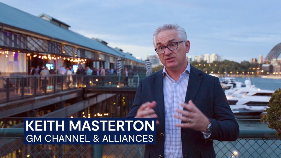 Person speaking on a waterfront boardwalk with marina and restaurant lights; on-screen text ‘Keith Masterton, GM Channel & Alliances.