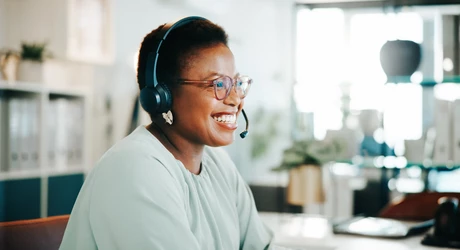 Person wearing a headset seated at a desk in an office, with a computer and shelves in the background. 