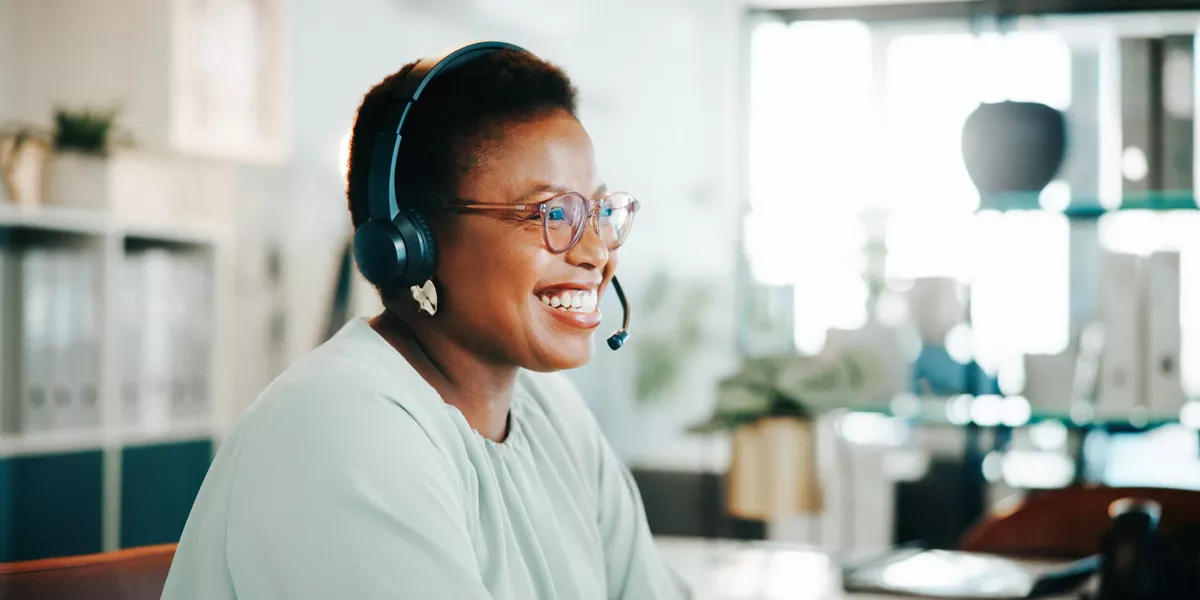 Person wearing a headset seated at a desk in an office, with a computer and shelves in the background. 