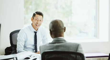 Two professionals having a one-on-one discussion across a desk in a bright office meeting room.
