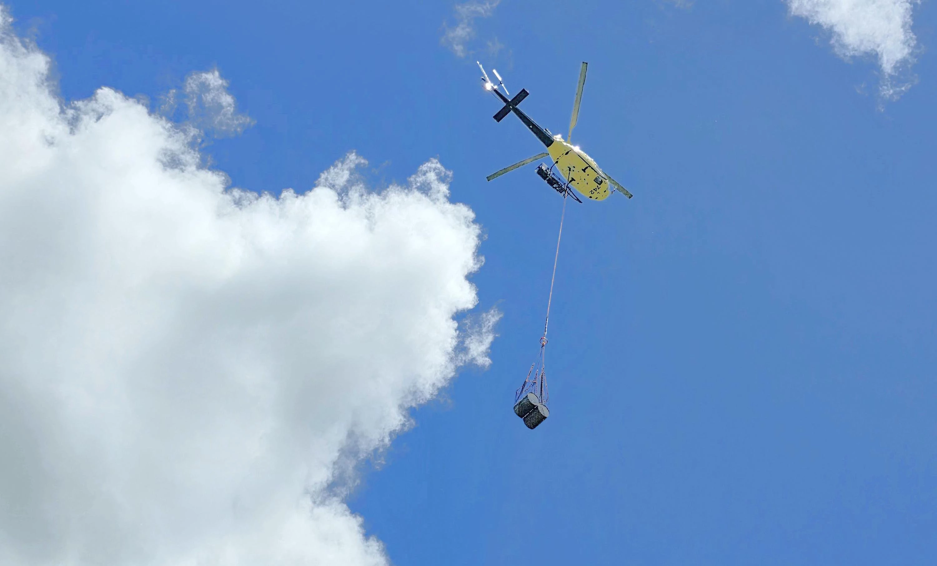 A blue sky and clouds with barrels of diesel being carried in a mesh sling underneath a helicopter connected by a cargo line