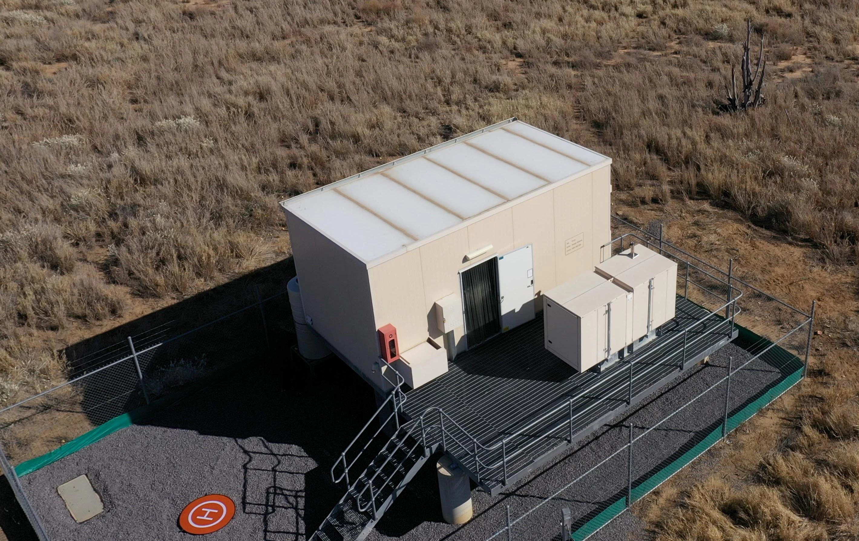 A building that looks a bit like a shipping container with a generator next to it on a gravel rectangle surrounded by cyclone fencing. The surrounds are low scrub typical of the Australian outback.