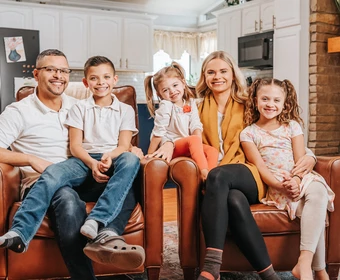 Family of five people seated together on chairs in a living room, with a kitchen, refrigerator, and fireplace visible behind them. 