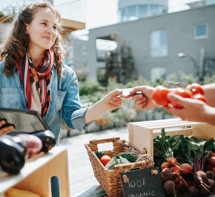 Woman using a tablet to complete a transaction at an outdoor market stand, while another person hands over produce, with baskets of vegetables on display. 