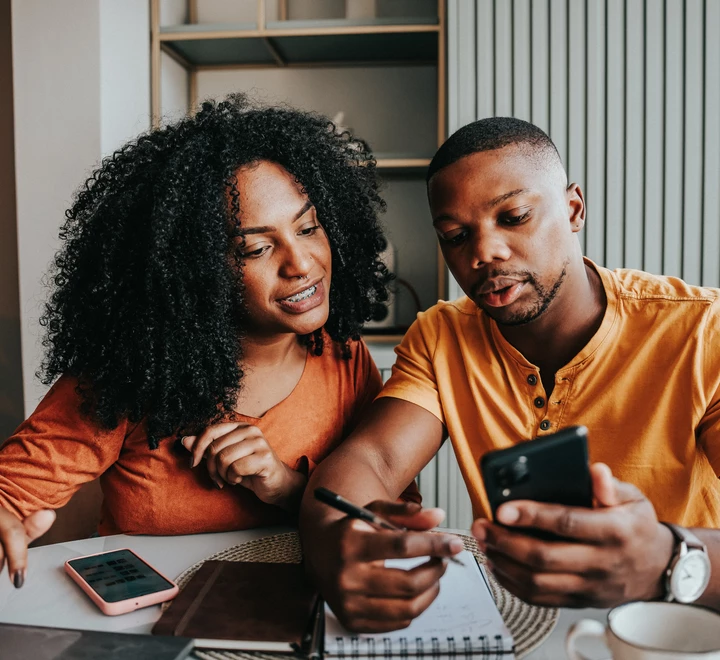 Two people sitting at a table indoors, looking at a smartphone together while one writes in a notebook 