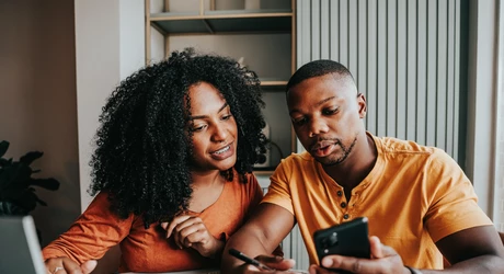 Two people sitting at a table indoors, looking at a smartphone together while one writes in a notebook 