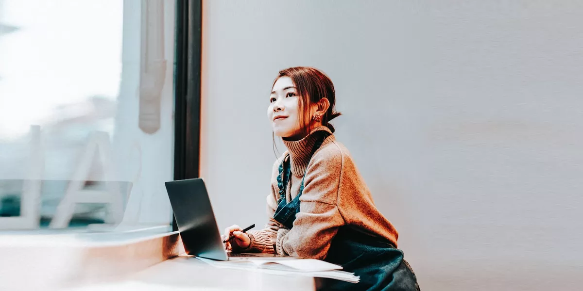 Person sitting at a desk with a laptop, looking out a window in a bright office.