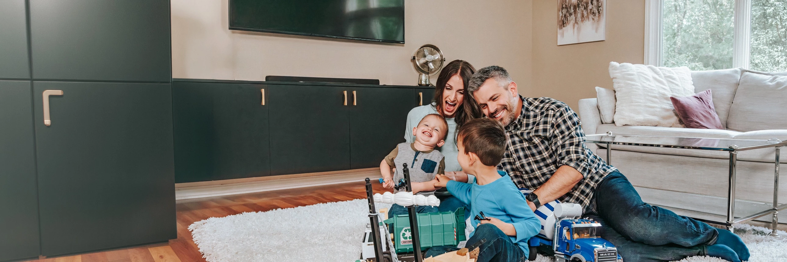 Two adult LMCU members and two children sit on a living room rug playing with toy trucks and construction vehicles.