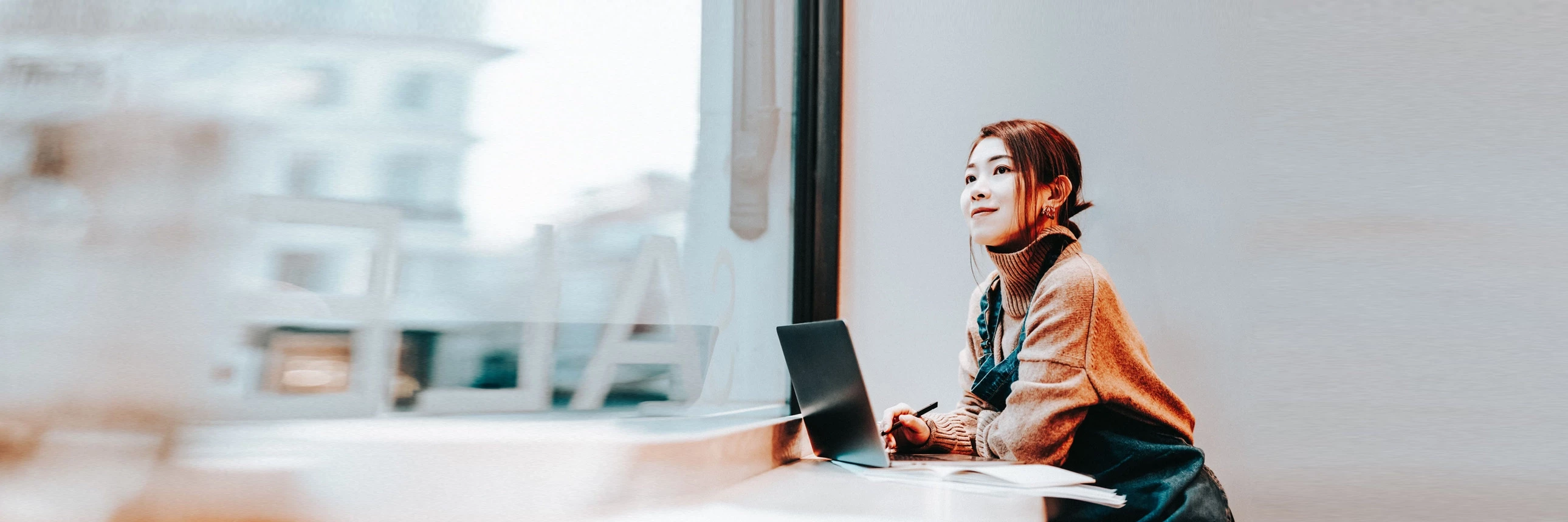 Person sitting at a desk with a laptop, looking out a window in a bright office.