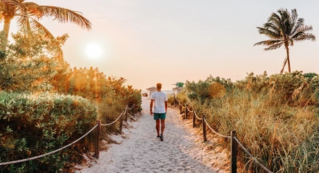 Person walking along a sandy path toward the beach at sunrise, surrounded by palm trees. 