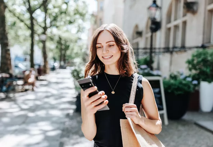 Woman walking outdoors on a street while looking at a smartphone, carrying a shoulder bag, with buildings and trees in the background. 