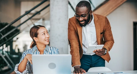 Man standing next to seated female coworker looking at laptop in office 