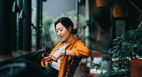Person sitting on a chair indoors, holding a smartphone, with potted plants and large windows in the background. 