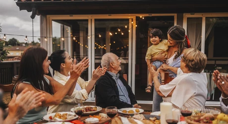 Multi-generational family in a backyard outdoor setting, eating together. The family is focusing on a young child that is being held by a female. Family is clapping. 