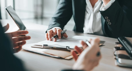 Business person's hand gesturing to clipboard on office desk 