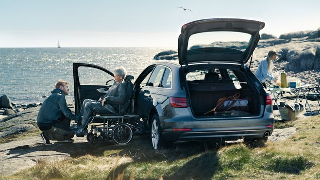 A man assists an older person in a wheelchair next to a silver car parked by the sea. The car's doors and boot are open, and another person prepares a picnic in the background. The scene is calm and scenic, set against a coastal backdrop.