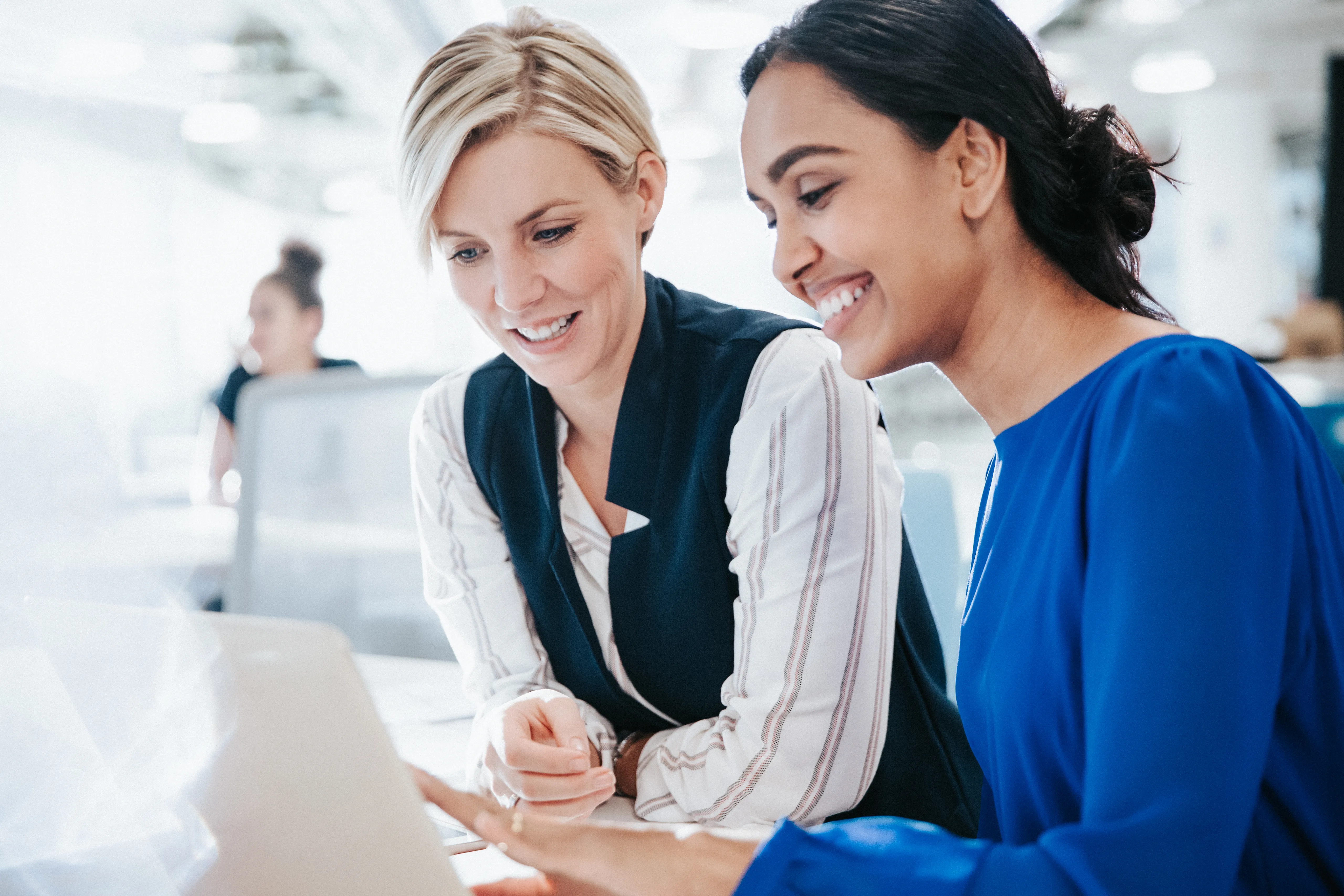 Two professionals in a bright office collaborate at a laptop, leaning in side-by-side while reviewing content on the screen.    