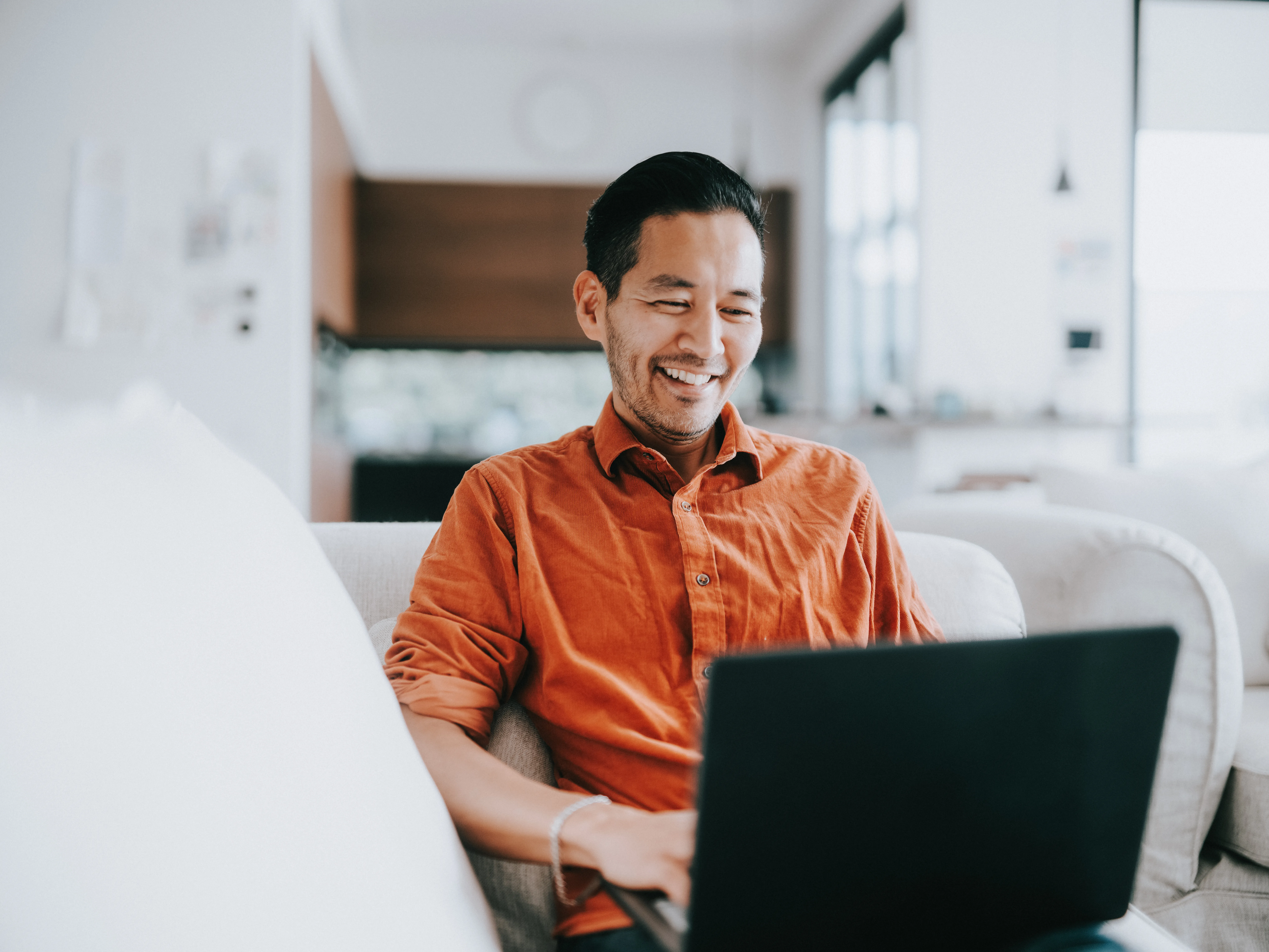 Male at home on a laptop, sitting on a couch, in a living room setting.  