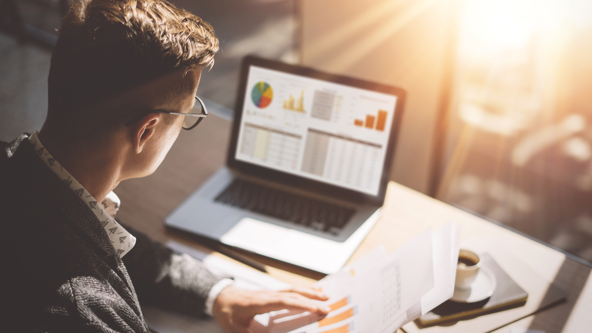 Young finance market analyst in eyeglasses working at sunny office on laptop while sitting at wooden table.Businessman analyze document in his hands.Graphs and diagramm on notebook screen.Blurred.