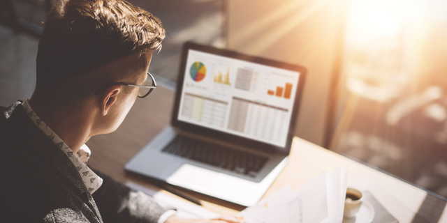 Young finance market analyst in eyeglasses working at sunny office on laptop while sitting at wooden table.Businessman analyze document in his hands.Graphs and diagramm on notebook screen.Blurred.