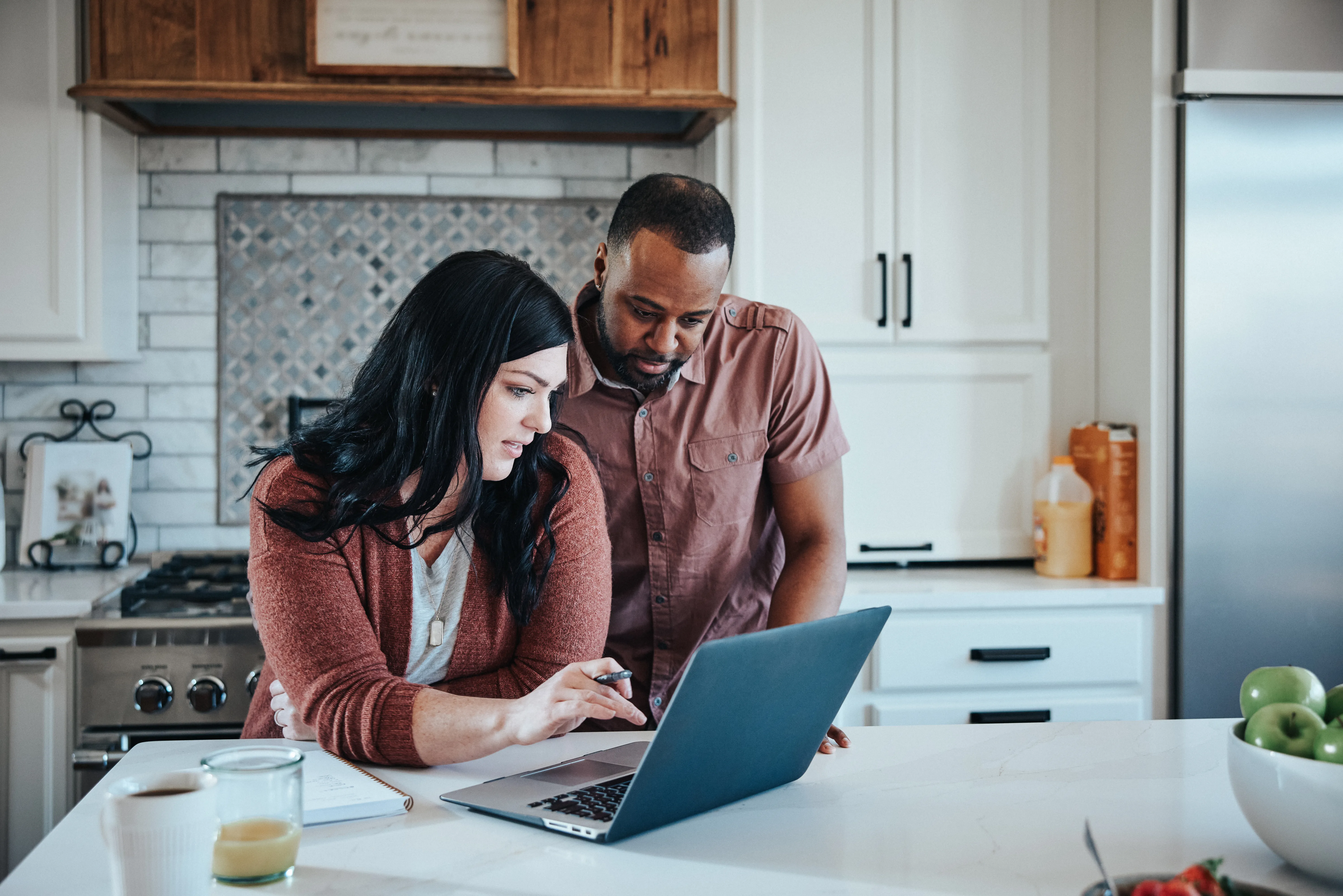 Man and woman sit at kitchen counter with breakfast working with pen, paper and laptop.    