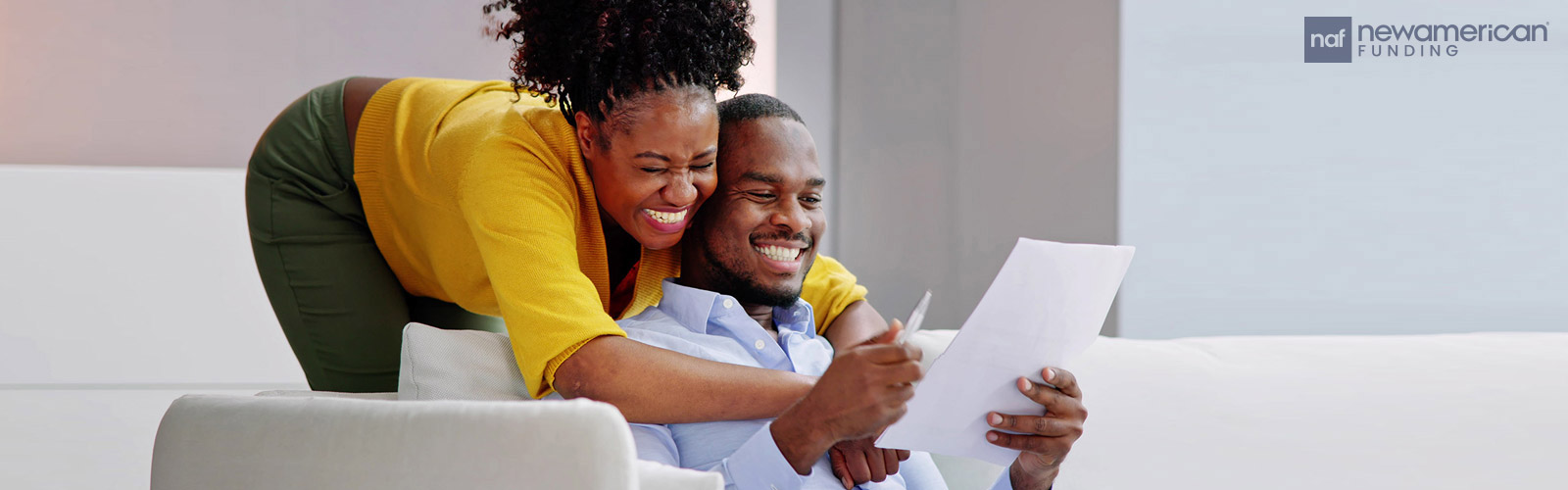 A Black man sits on a couch holding paperwork and smiling while a smiling Black woman wraps her arms around him