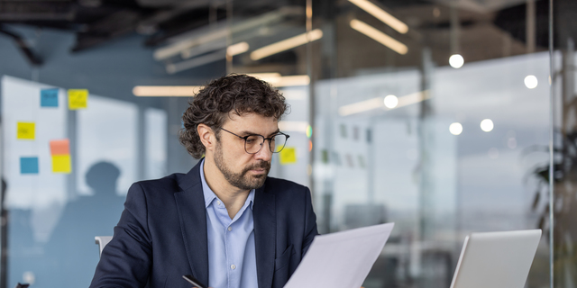 Serious businessman analyzing documents at office desk with laptop and papers