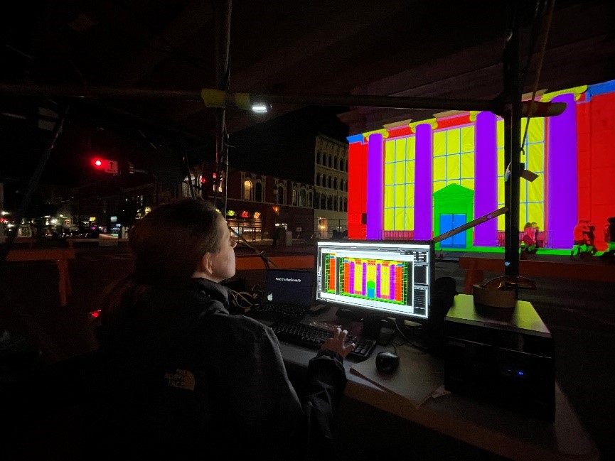 An AV technician sits behind the computer controlling a projection mapping display on a city building.