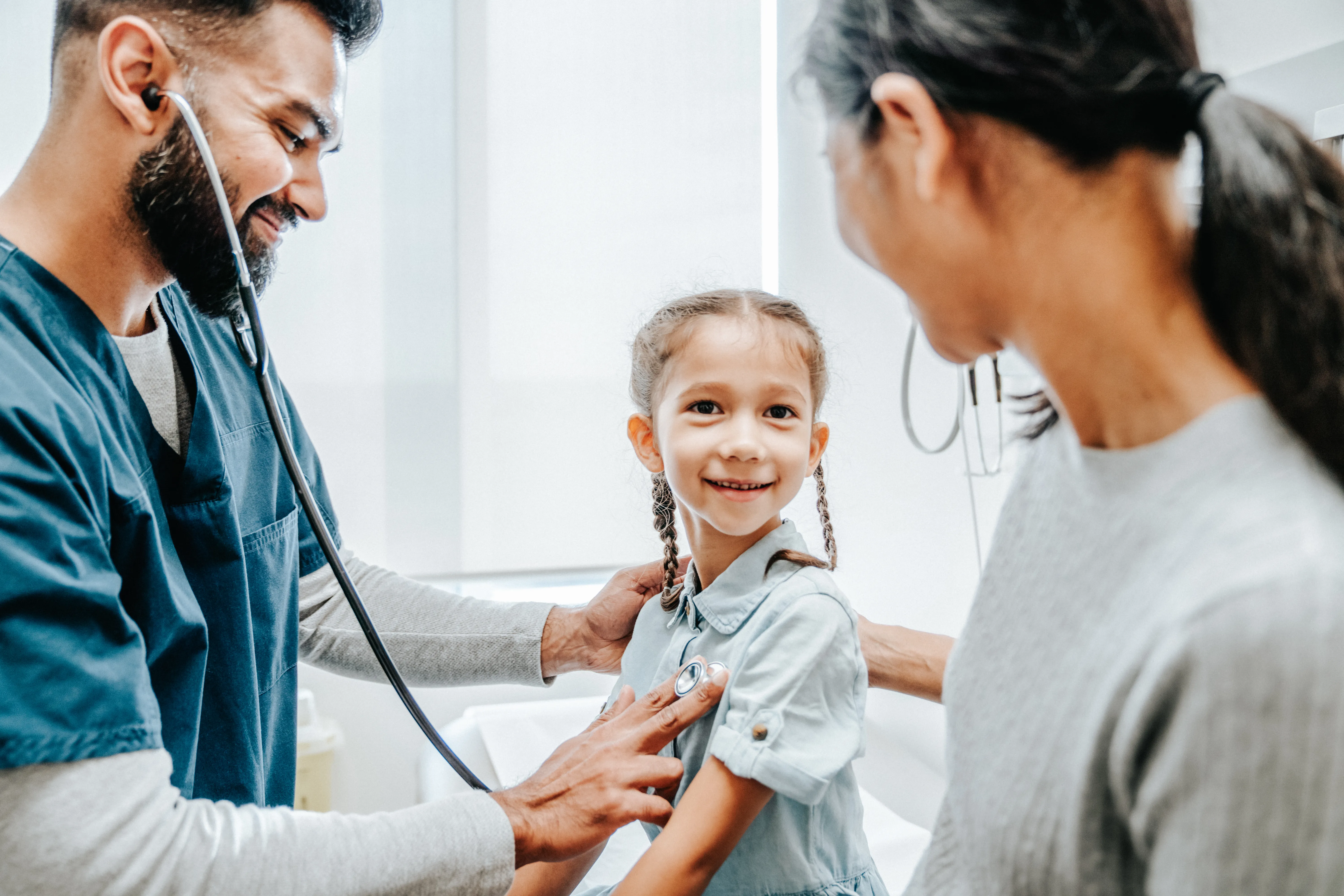 Male pediatrician listening to the heart of smiling young girl while her mother stands by 
