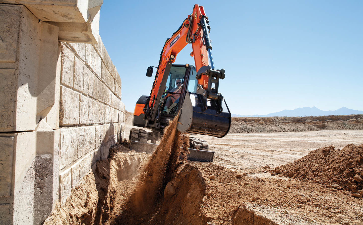 A DEVELON mini excavator digging next to a retaining wall on a job site.