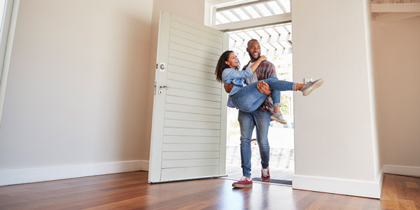 A Black man carries a smiling Black woman across the threshold of a new home