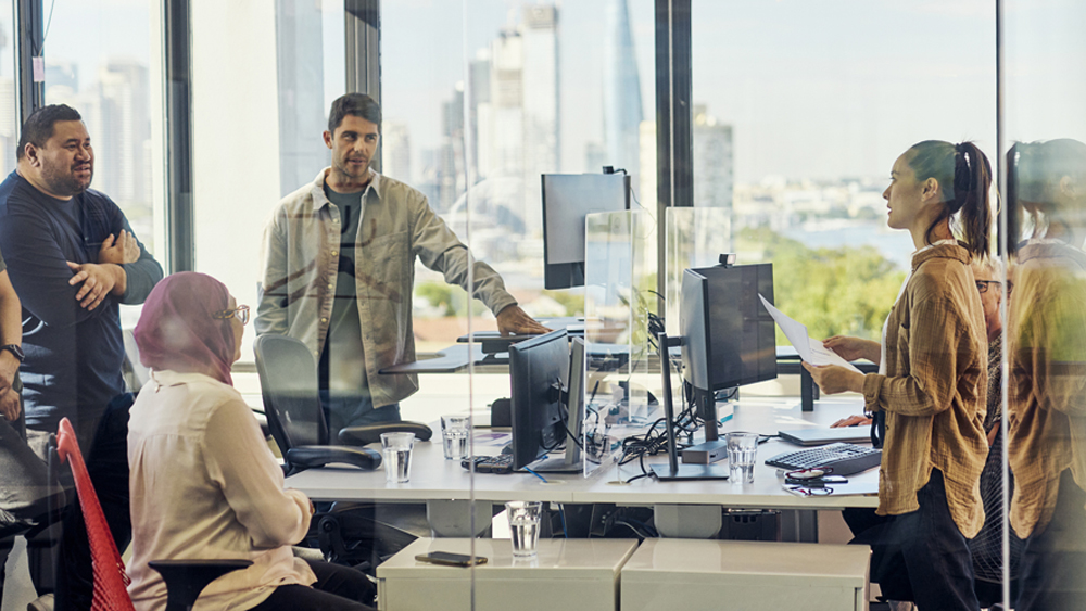 A group of people are gathered in a modern office space with large windows and a view of the city skyline. They are engaged in a discussion around a desk with multiple computer monitors, papers, and glasses of water. Some individuals are standing while others are seated. The room is well-lit with natural light coming through the windows.
