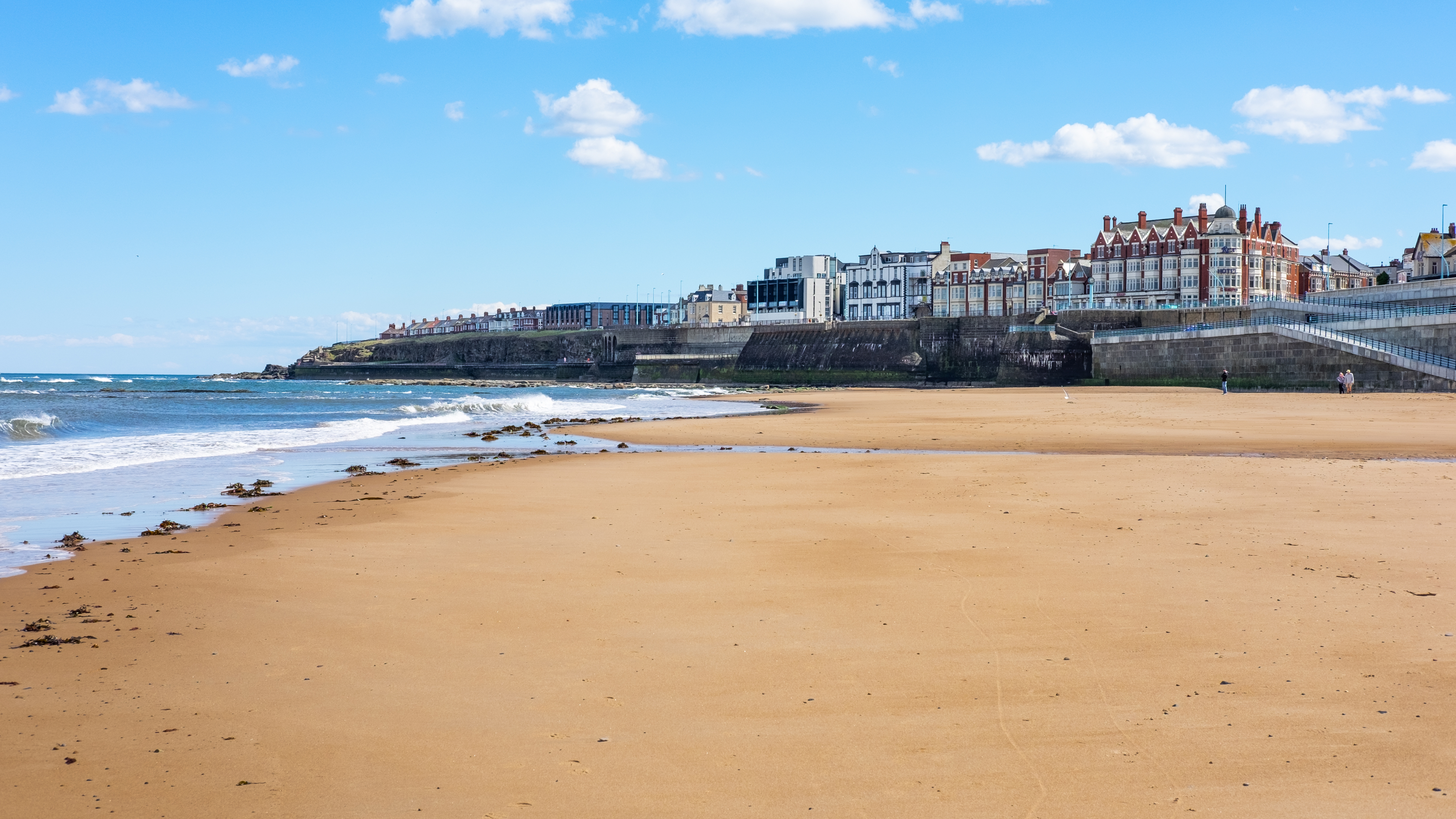 A view of Whitley Bay beach with golden sand and gentle waves. The town’s historic buildings and sea wall line the background, while a few people walk along the shore under a clear blue sky.
