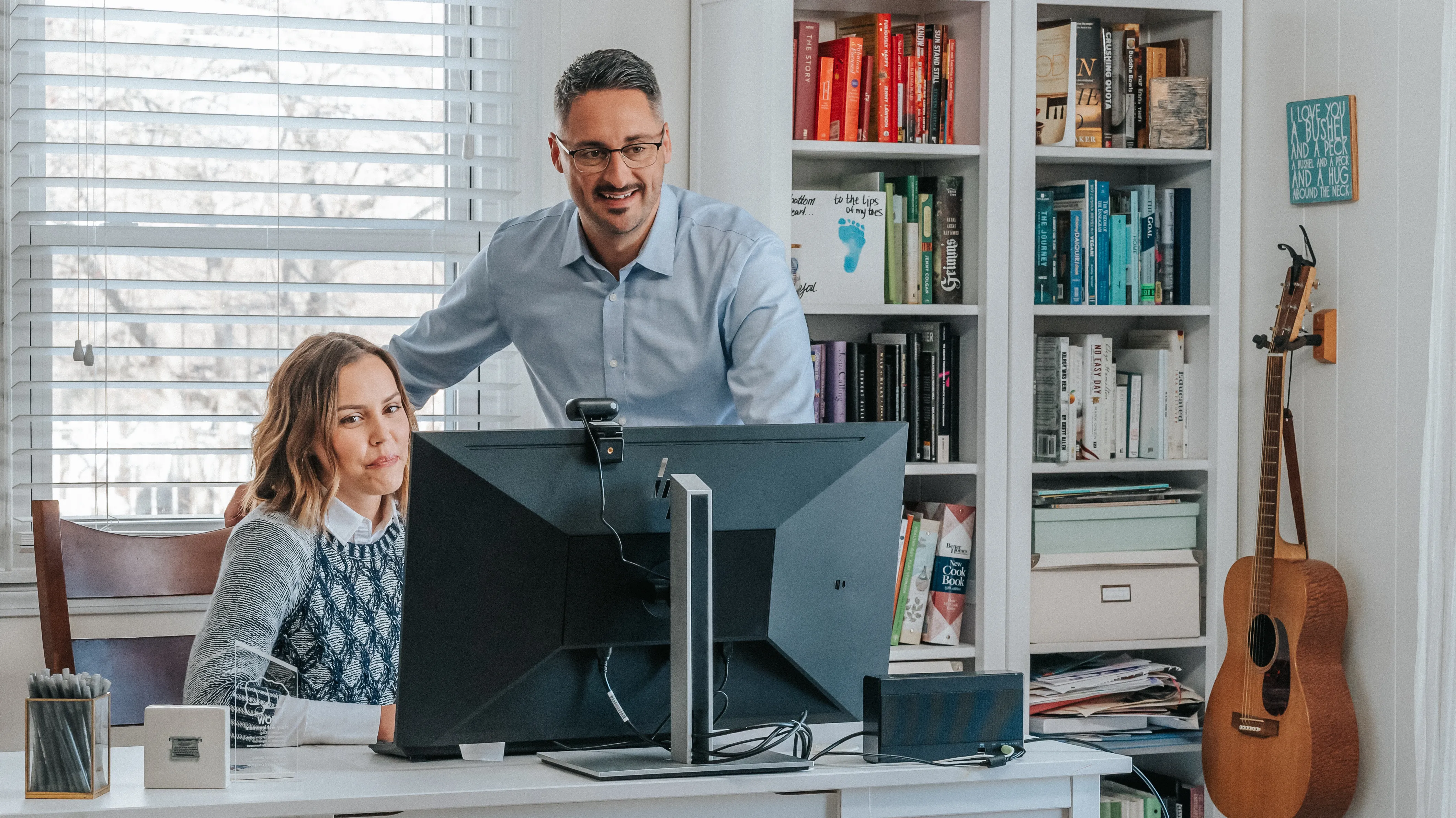 Two adults at a desk working on a computer in a bright home office, with one person seated and the other standing nearby. Behind them are bookshelves filled with books and decor, and a guitar rests against the wall next to the shelving. 