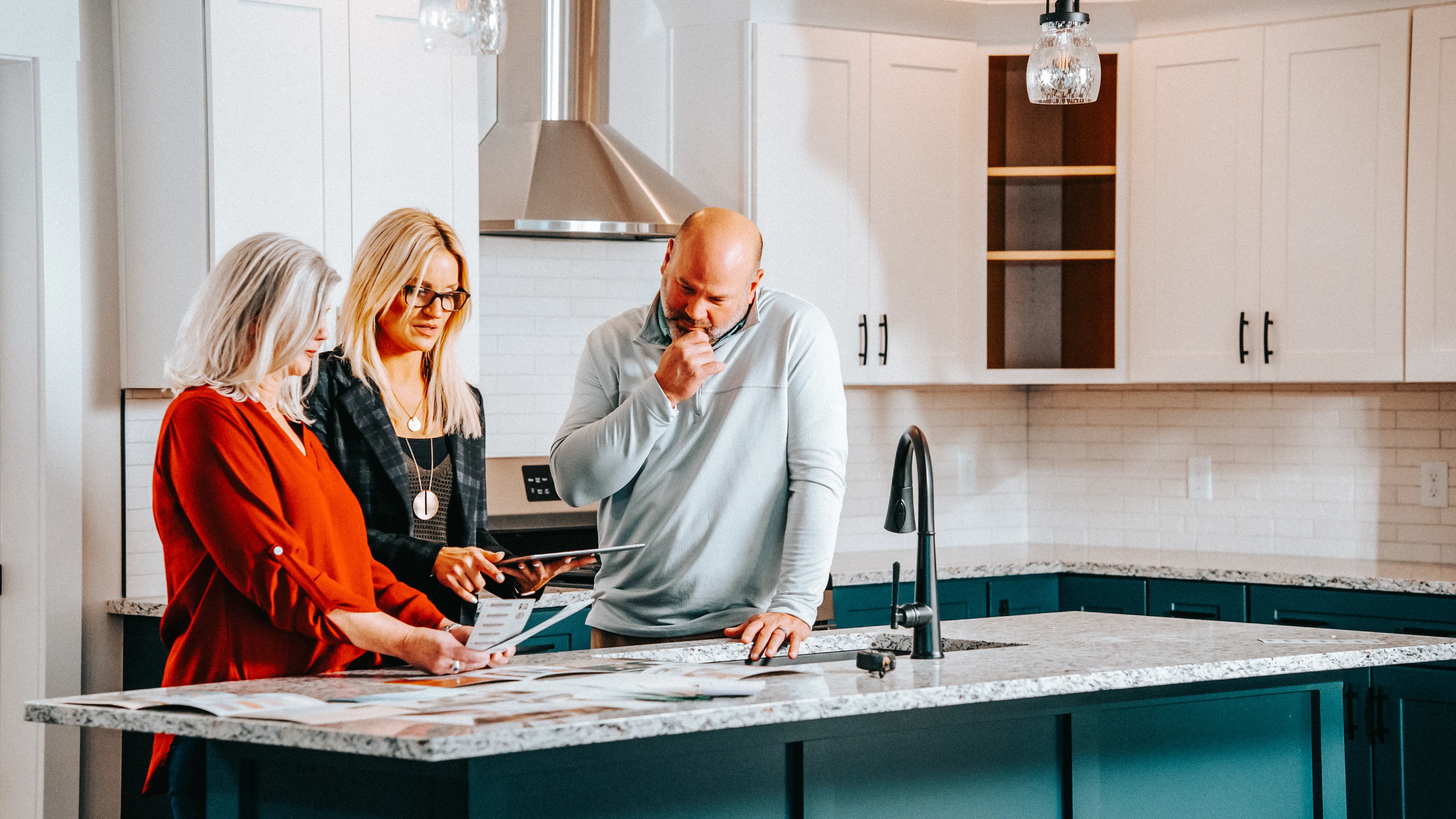 Three adults stand around a kitchen island reviewing a tablet in a bright, modern kitchen with white cabinets, pendant lighting, and a stone countertop. 