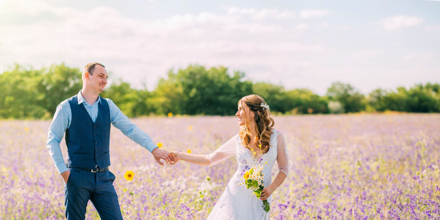 married couple walking in the field of purple flowers, the bride leads the groom behind her, the girl holds the guy by the hand, smiles, smoking hair, a woman in a white wedding dress, blue suit
