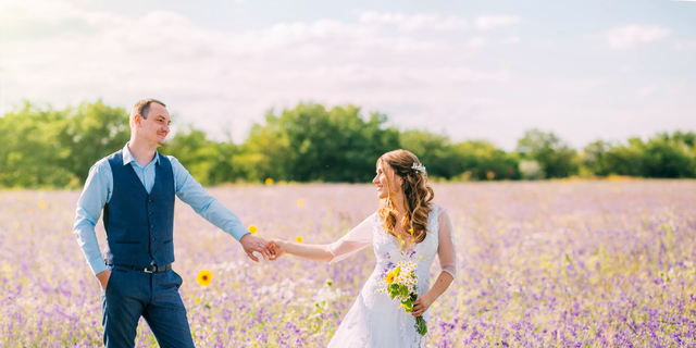 married couple walking in the field of purple flowers, the bride leads the groom behind her, the girl holds the guy by the hand, smiles, smoking hair, a woman in a white wedding dress, blue suit