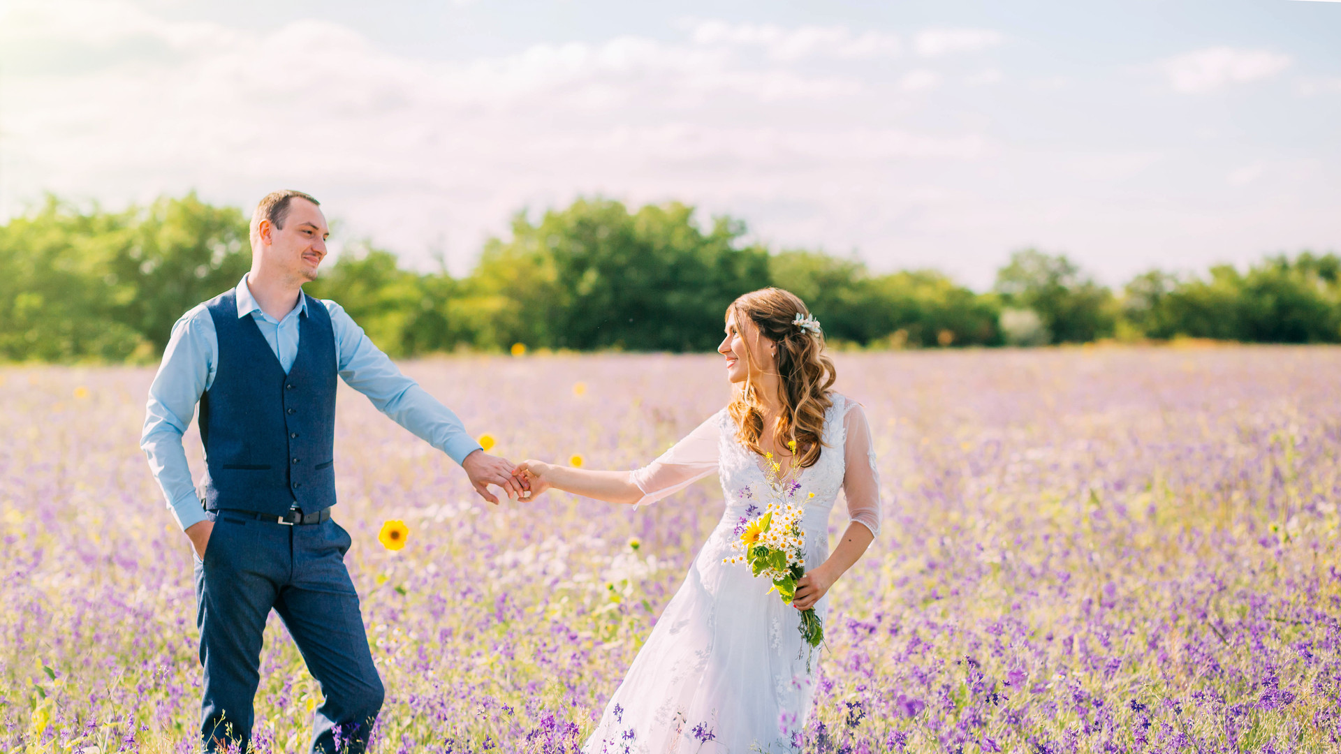 married couple walking in the field of purple flowers, the bride leads the groom behind her, the girl holds the guy by the hand, smiles, smoking hair, a woman in a white wedding dress, blue suit