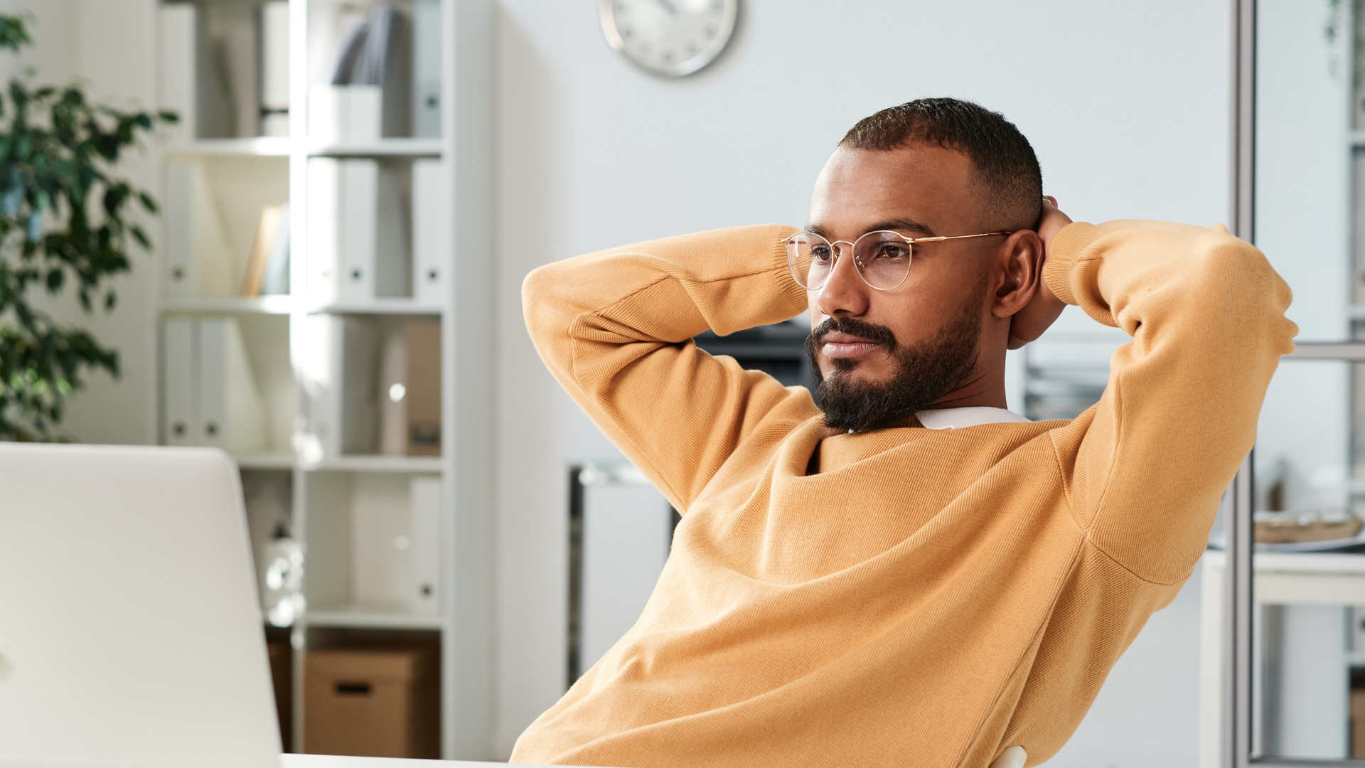 Serious pensive young black man in yellow sweater and eyeglasses sitting with hands behind head and looking at laptop screen in office