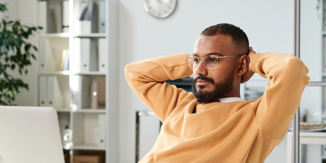 Serious pensive young black man in yellow sweater and eyeglasses sitting with hands behind head and looking at laptop screen in office