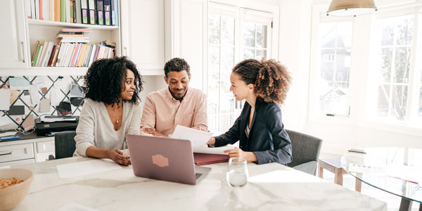 A Black man and a Black woman sit at a table covered with paperwork looking at a laptop screen with a Black woman wearing a suit jacket