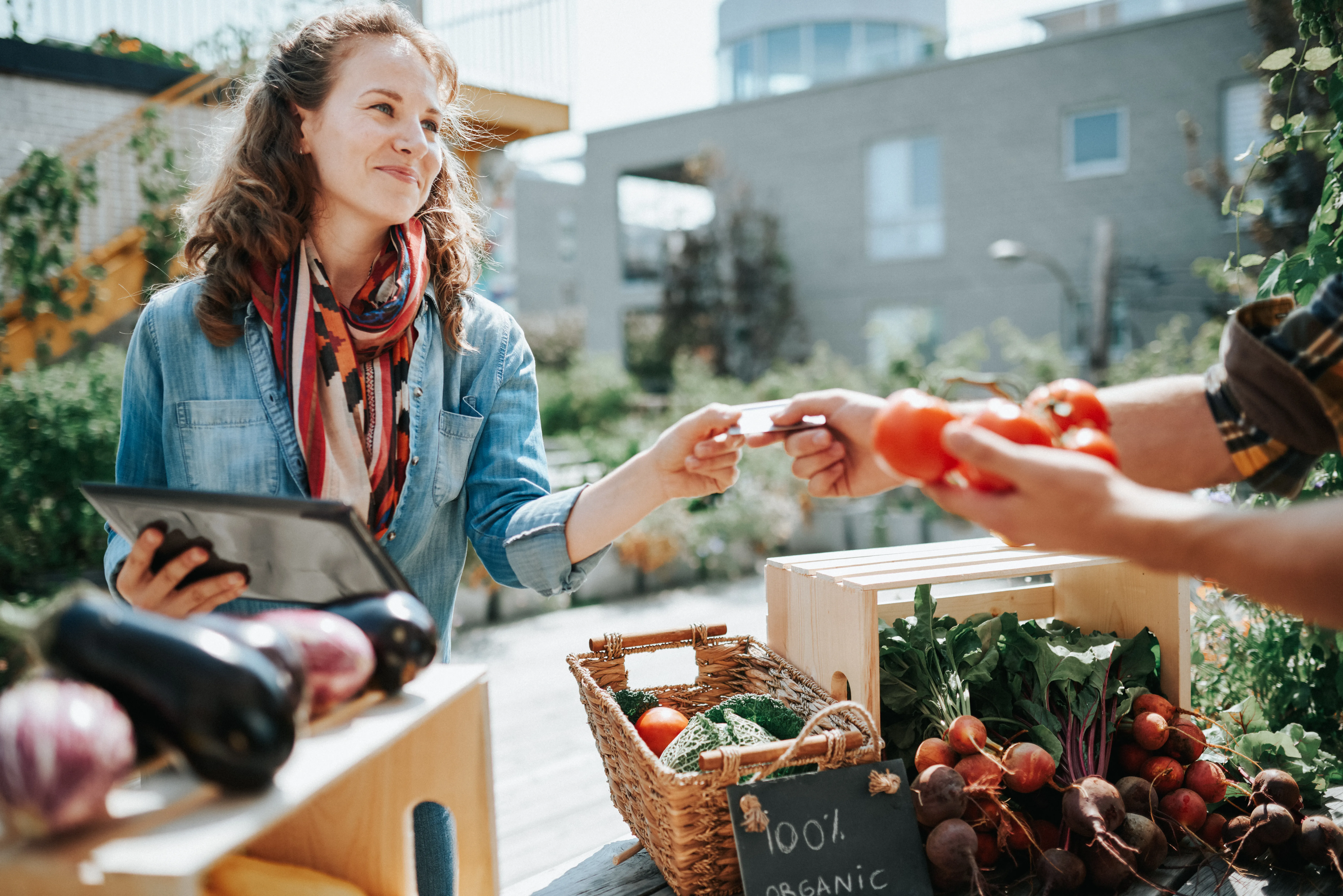 Woman using a tablet to complete a transaction at an outdoor market stand, while another person hands over produce, with baskets of vegetables on display. 
