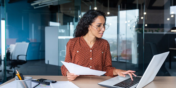 A woman sits in a office with papers in one hand typing on a laptop that is sitting on the table