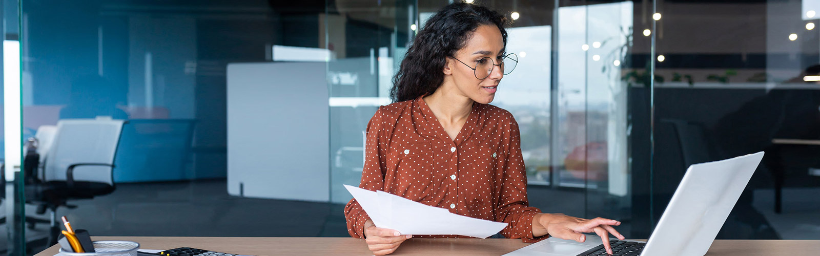 A woman sits in a office with papers in one hand typing on a laptop that is sitting on the table